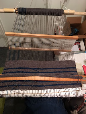 A short length of grey and teal warp threads stretched between a pair of pale wooden dowels with thread wrapped thickly around them, suspended by white cords at the ends that disappear out of frame at the top of the photo, and horizontally striped grey and teal cloth beginning to wrap around something at the photographer's lap. The heddle hangs in the middle of the warp threads, and a stick shuttle filled with grey yarn rests on the woven cloth.