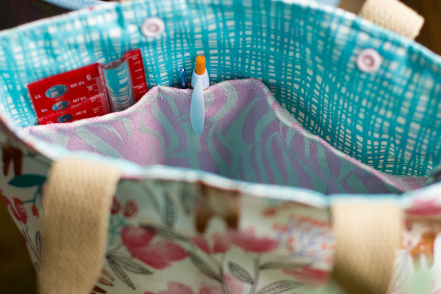 Interior view of a sloth patterned project bag with a teal hatchmark lining and pink and ecru wave patterned triple pocket. Pocket holds a red ruler and a pen and crochet hook.