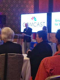 Man standing in front of podium with a lare screen in the background that says comcast. Also two women sitting at  table listening to presenter