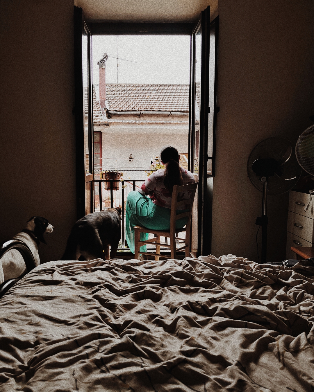 A woman is sitting on a chair facing the balcony in her bedroom and looking out at the street. Behind is the bed and next to her are her two dogs who are about to go on the balcony too