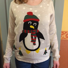 A light grey jumper, with a giant penguin wearing a red and green woolly hat and scarf, in a snow scene, with tiny Christmas trees.