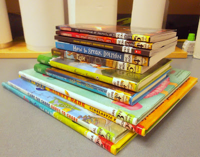 Stack of books, their spines visible with library spine labeling. Behind the stack are vertical rolls of protective dust-jacket covering.