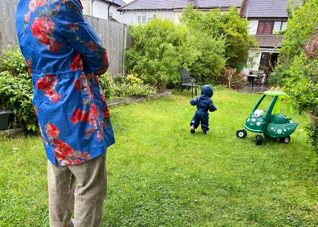 A toddler in a wet garden in a navy puddlesuit at a socially distanced 2 metres from an adult in a waterproof to stay dry from the rain