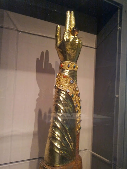 Golden hand and arm in an upraised position, a large jeweled ring on the hand, a bracelet of gems on the wrist, and a bejeweled band wrapped diagonally around the forearm