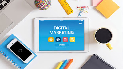 White on blue digital marketing sign projected from tablet screen on a table and surrounded by a smartphone on top of a notepad, pens and pencils, paper clips, ruler, notepads and a white mug of coffee.