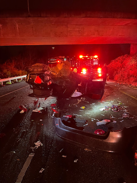 : A wrecked car lies on the side of the road in the dark with an EMS vehicle staged in the background.