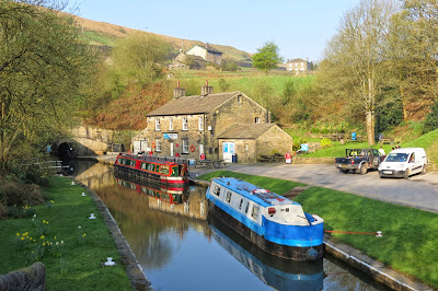 Two narrowboats moored by the stone arch of the tunnel entrance. A stone building stands to the right of the tunnel with hills and other buildings behind.