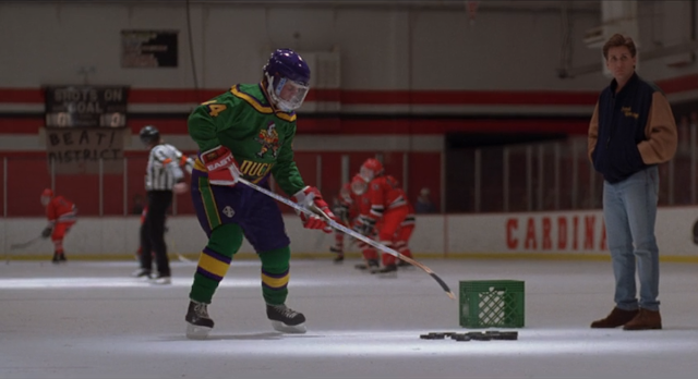 Gordon Bombay, interpretado por Emilio Estevez, treina os jogadores dos Mighty Ducks em uma quadra de patinação no gelo.