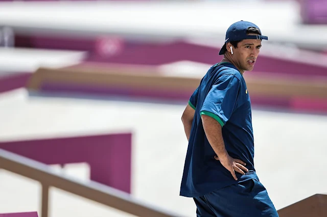 Kelvin Hoelfer de boné, camisa e shorts azul na pista de skate com as mãos na cintura e fone de ouvido