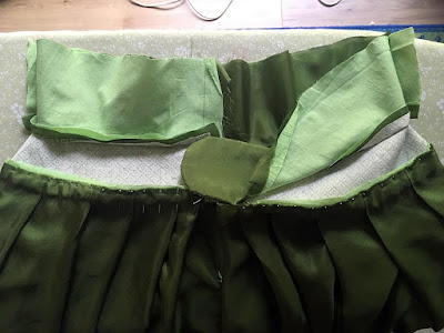 The guts of a green skirt's three-layered waistband, showing a clean-finished button tab and many, many pins attaching a deep-apple-green pleated layer to the lower edge of the bottommost layer.