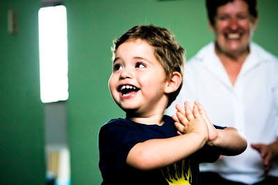 A fotografia mostra em primeiro plano um menino sorrindo com o rosto voltado para a sua direita e mãos espalmadas na altura do peito. Ele tem cabelos dourados e curtos e veste uma camisa preta com estampa frontal. Atrás dele, em desfoque, uma senhora de cabelo curtos e roupas brancas esbanja um sorriso ao olhar o menino. Ao fundo, uma parede verde-folha, com frestas iluminadas.