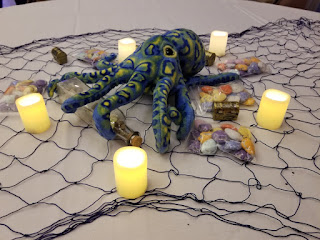 Table centerpiece with plushy Kraken, candles, a ship in a bottle, shells, annd treasure chests. They are sitting on netting.