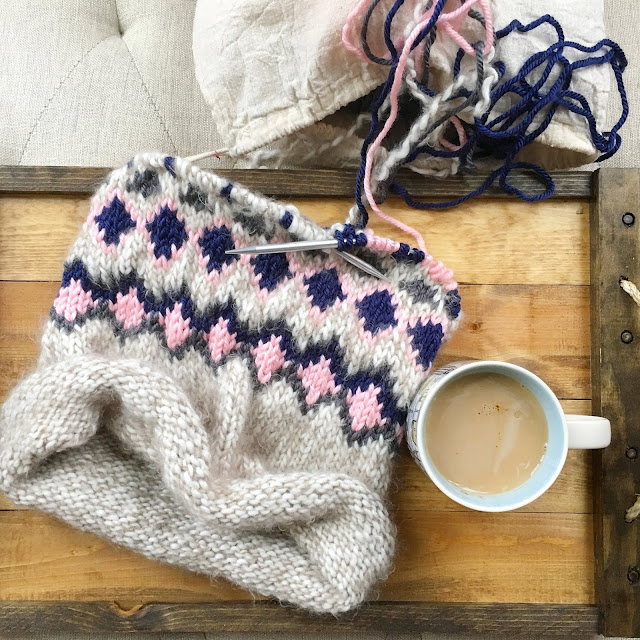 [Flat shot of a four color knit cowl on circular knitting needles next to a cup of coffee on a wooden tray. Colors of yarn include beige, light pink, charcoal gray, navy.]