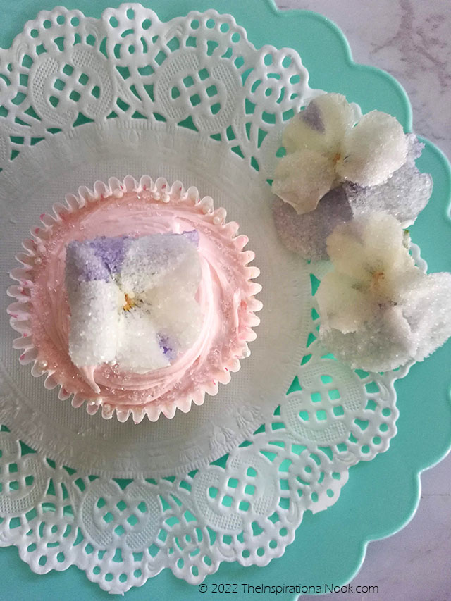 Pink iced cupcake with candied pansy flower on white paper doily and turquoise plate