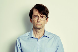 David Tennant looking serious with lank hair, big round glasses and a blue shirt with a large collar