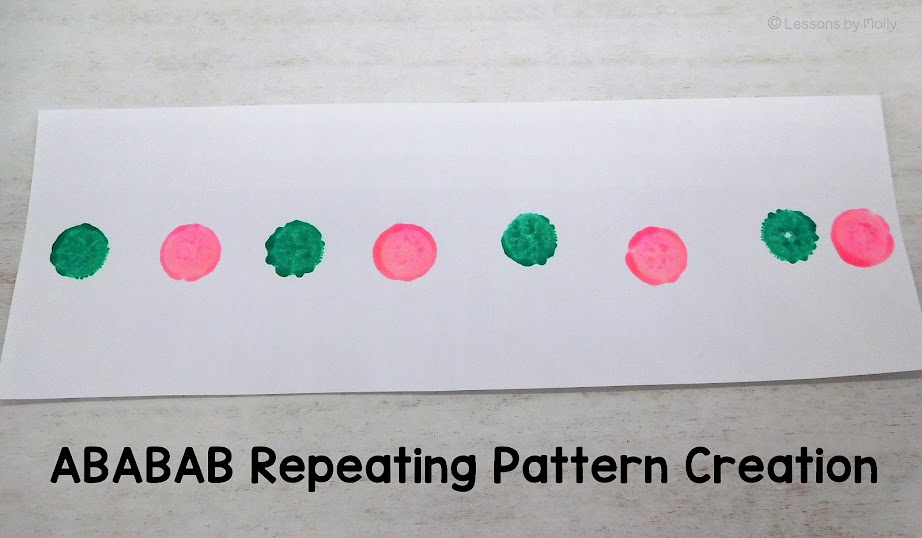 A strip of white construction paper is displayed, showcasing a creative expression of an ababab pattern using color. The pattern has been formed by a child (although not visible in the photo) using pink and teal paint daubers. The alternating sequence of pink and teal dots creates a visually appealing and clearly discernible pattern along the length of the paper strip.