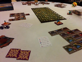A game of Between Two Cities in progress: the scoring board is in the centre, with the square tiles that form the cities in groups around the edge of the playing area. Stacks of unused tiles, marked with player pieces, sit nearby.