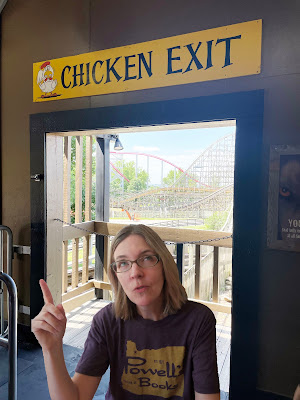 A light-skinned woman with blond hair in a dark grey shirt stands in a doorway with a rollercoaster in the background. Above the doorway is a sign that says "Chicken Exit."