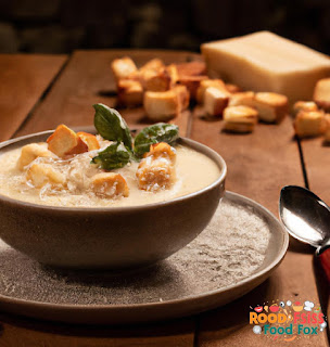 Image of the creamy soup in a bowl, topped with a few croutons, a sprinkle of parmesan cheese, and a leaf of fresh basil on top. The bowl is placed on a wooden table with a spoon next to it.