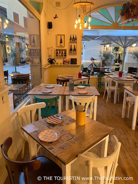 Five whitewashed square-sized bistro tables with wooden tops and a mix of green, white, and wooden bistro chairs in a bohemian-style decorated restaurant with two large shop windows, in the soft light of early evening.