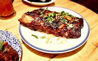 A half rack of dark brown pork ribs with cylindrical bones still attached with a dark brown Coca Cola glaze with red chilli pieces sprinkled on top on a white circular plate with a blue rim on a light golden brown table on a light background. 