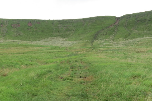 A path heads though grassland to the long ridge on the horizon, which it climbs steeply.