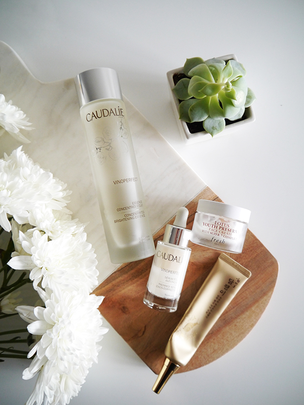 Flat lay featuring Caudalie Vinoperfect Concentrated Brightening Essence and Radiance Serum, Fresh Lotus Youth Preserve moisturizer, The Face Shop eye cream