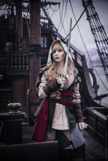 This woman is dressed as a female steampunk pirate (lady pirate). Clothing: hooded shirt, jacket, red sash, belt, boots. Women's steampunk pirate costume. wench/buncaneer