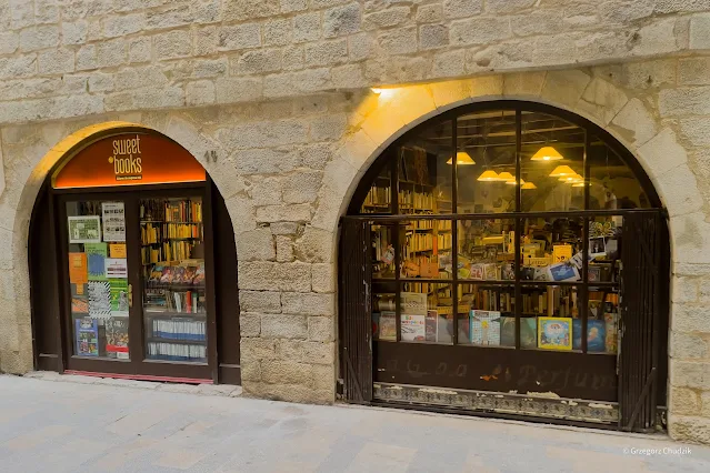Zewnętrzna fasada księgarni Sweet Books w Gironie, z dwoma łukowymi wejściami i witrynami pełnymi książek, osadzona w kamiennym murze historycznego budynku.