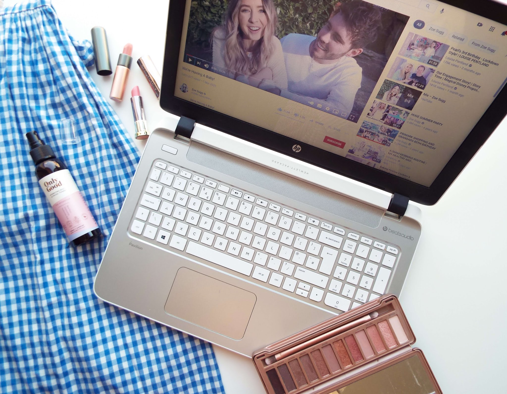 Flatlay of my March favourites, consisting of blue gingham dress diagonally to the left; skincare sat on top of the dress; my laptop centre, with Zoe Sugg's vlog displayed; some lipsticks to the top left area and eyeshadow palette to the bottom right.