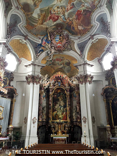 An elaborately decorated church interior with eight rows of dark wooden benches, white stone columns, and railings, an altar decked with candles and paintings, under a ceiling painting with a white and blue flag, a group of people playing instruments, painted in softened colours ranging from purple, pink, light blue, and red to gold.