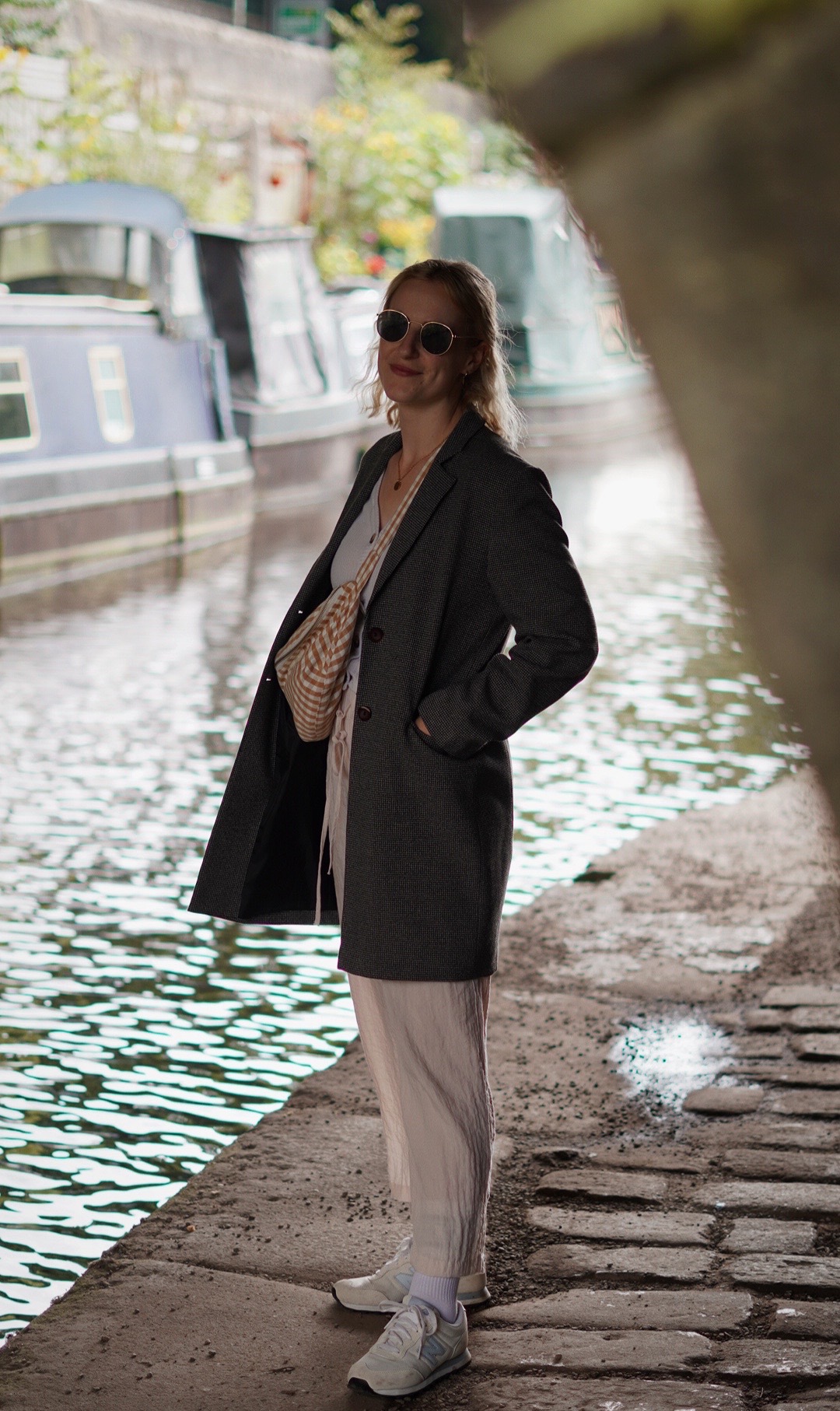 Amy is standing underneath a bridge next to a canal, looking to camera. She is wearing cream linen trouser and a grey long blazer