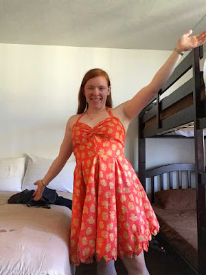 A young redheaded white woman in a room with a dark bunk bed and a low regular bed, posing with arms outstretched and wearing a bright coral halter-top dress with a full, knee-length, scallop-edged skirt.