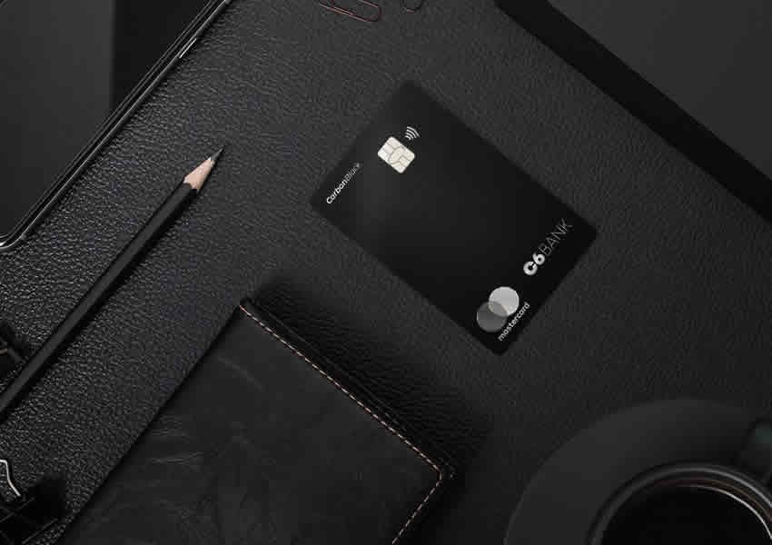 Imagem de cor escura em textura em couro com um cartão C6 Carbon sobre ela ao redor vários objetos como um lápis uma carteira e Smartphone.