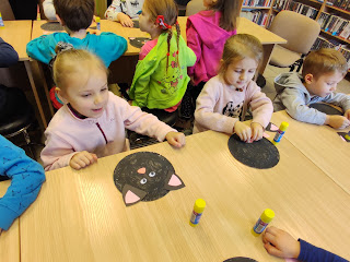 Dzieci wykonują pracę plastyczną - papierową mordkę kota. Tło: sala biblioteczna