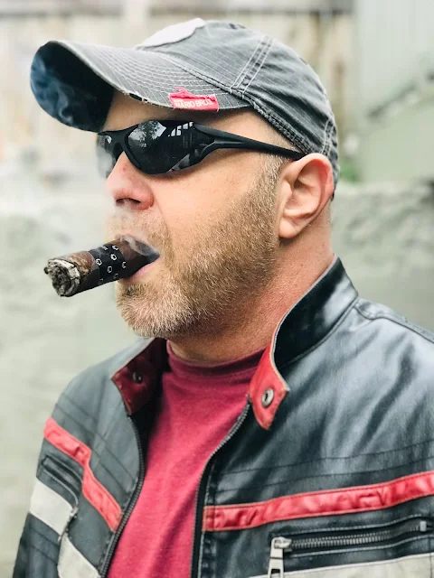 Baker with a cigar in his mouth wearing a baseball cap and a leather jacket that's red black and white with a red t-shirt underneath