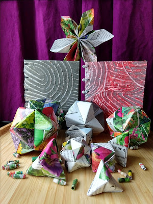 Background has pleated paper star on top of 2 paste-paper covered books with beaded spines. Foreground has a variety of modular origami polyhedra and some scattered, rolled paper beads.