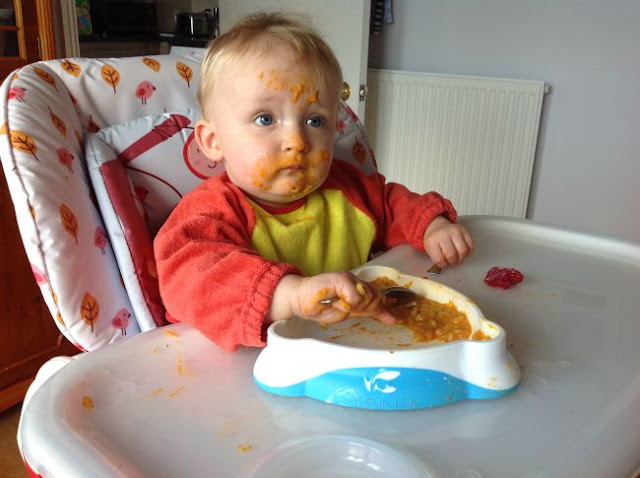 baby in highchair with spoon in hand and face covered in food