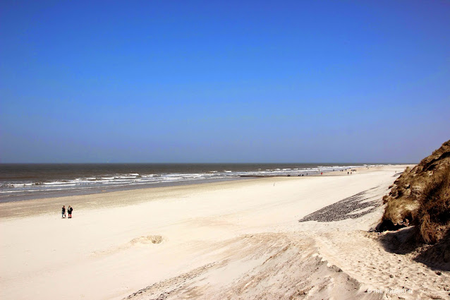 Two people, under a big blue sky, walking on a white endlessly long sandy beach.