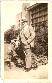 man in business suit hat glasses standing in front of multi story building in 1930s