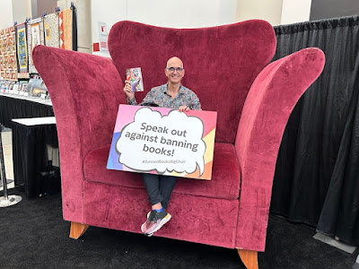 Lee Wind sitting in the very big chair at ALA Annual, with a sign reading "Speak Out Against Banning Books!"
