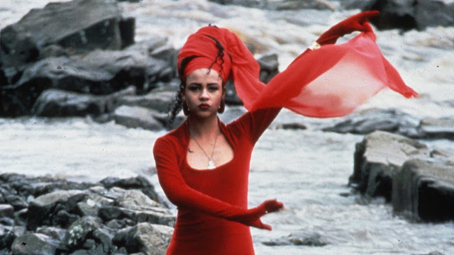 Film still from The Body Beautiful. A woman stands by a body of water in a red dress. Her arms are outstretched to the left.