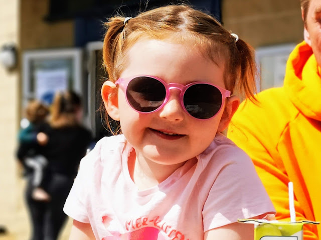 Image of a young girl wearing a pink t shirt and a pair of pink sunglasses. She is sat in the sunshine and is smiling broadly.