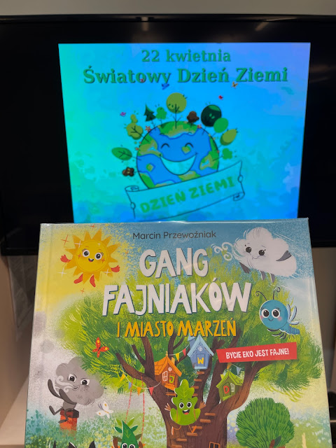 U góry slajdy tytułowy, wyświetlony na telewizorze. Na slajdzie napis 22 kwietnia Światowy Dzień Ziemi oraz grafika, przestawiająca uśmiechniętą Ziemię. Poniżej okładka książki "Gang Fajniaków i miasto marzeń", autorstwa Marcina Przewoźniaka.