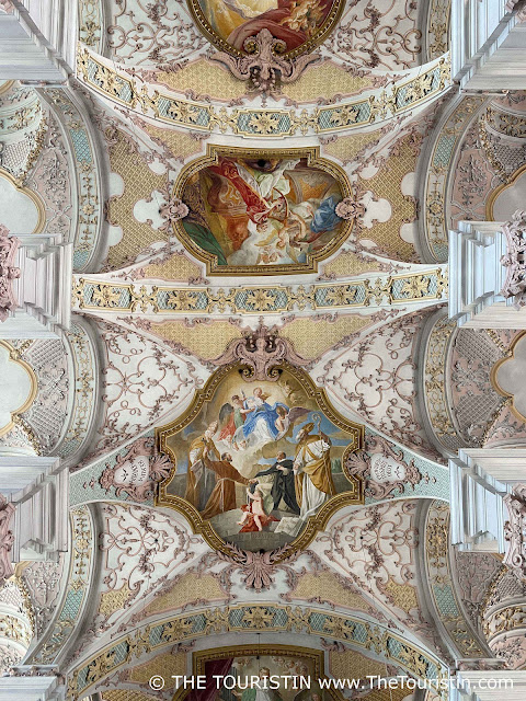 An elaborately decorated ceiling painting with a groups of people dressed differently in coloured robes and tunics, painted in pastel colours ranging from pink, light blue, and yellow, to red to gold.