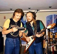 Rudolf e Michael em 1985 em estúdio. Rudolf segura algo que parece um gravador e Michael, com fone de ouvido ligado ao gravador, toca sua guitarra flying V.