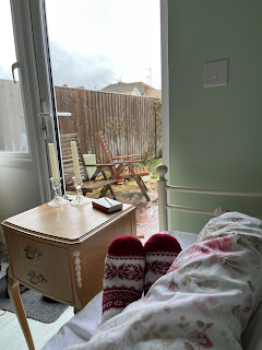 The fluffy-socked feet of someone in bed, behind which are a bedside table with 2 candles on, and a glass door through which a rainy patio, some garden furniture, and a few plants are visible.