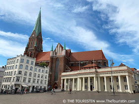 A market square with only a few pedestrians framed by white-pillared property and a large red-brick cathedral under a blue sky.