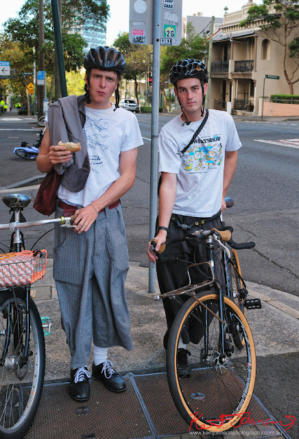 Two men with long baggy pants and cool bike walking on a footpath eating sandwiches