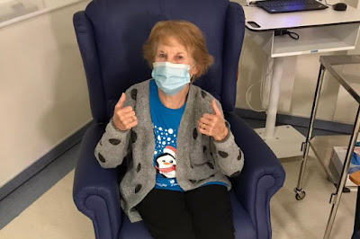 Margaret Keenan First UK patient vaccinated against COVID Pfizer BioNTech, sitting in the vaccination chair double thumbs up